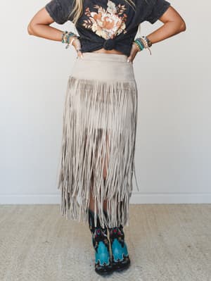 A back-facing model styled in a knotted floral graphic tee and long suede fringe skirt in dove, paired with embroidered turquoise cowboy boots and stacked boho bracelets — the kind of outfit that has a whole personality.