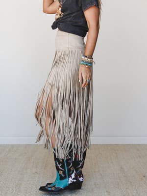 A side-profile shot of a model in a long suede fringe skirt in dove cream, knotted graphic tee, embroidered cowboy boots, and a stacked arm of beaded bracelets — the kind of outfit that has a road trip playlist to go with it.