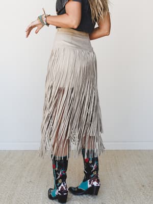 Shot from behind, a model in a faded black tee and embroidered cowboy boots wears a long suede fringe skirt in dove — all movement and texture, like she's already halfway to the dance floor.