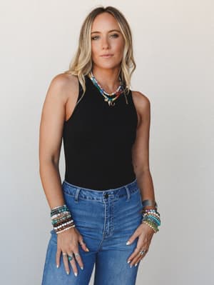 A relaxed-cool studio shot of a model in a fitted black sleeveless bodysuit and high-rise blue jeans, styled with layers of colorful beaded necklaces and an arm full of stacked bracelets — the kind of outfit that looks effortless but clearly had thou