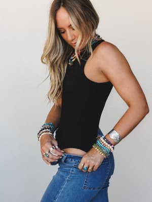 A model in a fitted black racerback bodysuit and medium-wash jeans, styled with stacked beaded bracelets, a silver cuff, layered necklaces, and rings — the kind of effortless accessory moment that makes the simplest outfit feel completely personal.