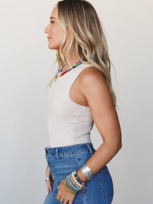 A side-profile shot of a model in a fitted sand beige sleeveless bodysuit tucked into medium-wash jeans, styled with stacked colorful beaded bracelets, a silver cuff, and a multicolor beaded necklace — the kind of effortless everyday look that does a
