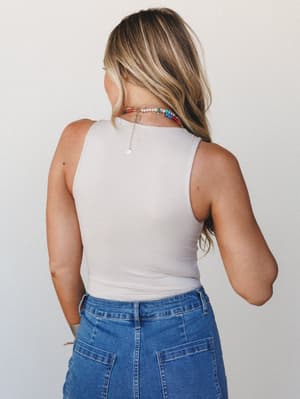 Shot from behind, a model in a fitted sand beige sleeveless bodysuit and medium-wash denim skirt layers on colorful beaded necklaces — casual, effortless, the kind of outfit that looks good without trying.