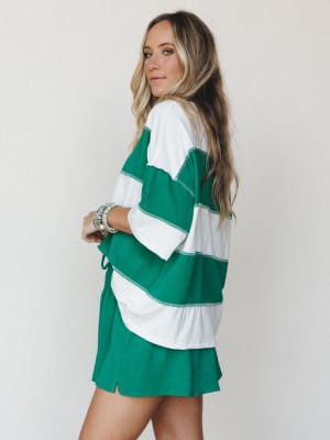 A model shot from behind at a three-quarter angle showing a coordinated two-piece set — an oversized wide-stripe top paired with matching solid shorts — in kelly green and white, styled with stacked beaded bracelets against a soft neutral studio back