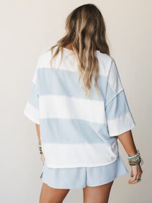Shot from behind, she's wearing a relaxed oversized striped top and matching shorts in soft sky blue and white — stacked beaded bracelets on both wrists adding just the right amount of effortless personality.