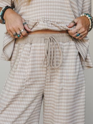 A close-up detail shot of a matching gingham pant set in soft sand and cream, styled with an armful of silver and turquoise jewelry — the kind of outfit that feels like a Saturday morning done right.