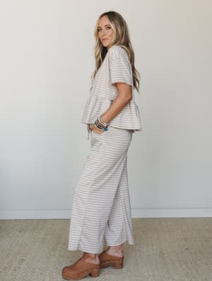 A matching gingham set in warm sand tones — peplum top and wide-leg pants styled with wood-studded clogs and a stack of beaded bracelets, shot against a clean white studio wall with an easy, lived-in confidence.