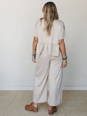 Shot from behind, a model wears a matching gingham pant set in warm sand — a peplum top and wide-leg cropped pants — styled with brown platform clogs and stacked turquoise and beaded bracelets for that effortless, collected look.