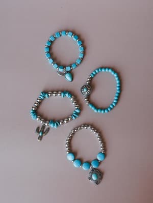 Four turquoise and silver stretch bracelets laid out on a dusty mauve surface — each one slightly different, with stone beads, chip beads, silver balls, and western-inspired charms like a cactus, sunburst, and oval concho pendant.