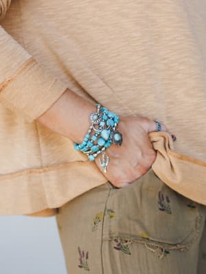 A close-up of a stacked turquoise bead bracelet set layered on the wrist, styled against a sandy camel top and olive floral-embroidered pants — earthy, expressive, and very much the kind of arm party that starts conversations.