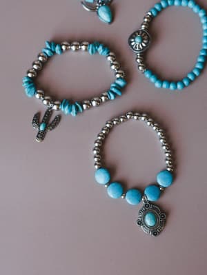 A flat lay of four turquoise and silver beaded stretch bracelets scattered on a dusty mauve surface — each one a little different, with details like a cactus charm, a concho pendant, sun medallion, and raw chip stones that feel like they belong toget