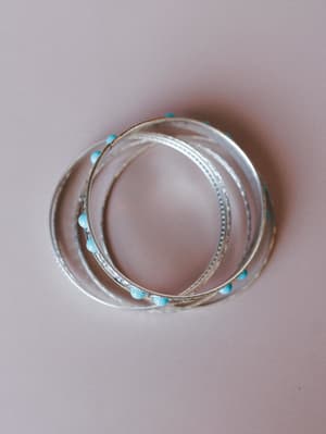 A stacked set of silver bangle bracelets with turquoise beads dotted along the band, layered together and shot from above on a soft blush surface — the kind of arm candy that goes with everything and somehow makes the whole outfit.