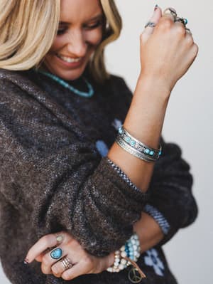 A smiling woman in a chunky dark knit sweater shows off a stacked mix of silver and turquoise jewelry — bangles, rings, and beaded bracelets layered with easy, collected intention.