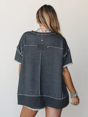 A back-view shot of a relaxed charcoal short set with exposed seam detailing and raw edges — the kind of set that looks like you tried just enough, styled with stacked boho bracelets and a delicate necklace peeking out.