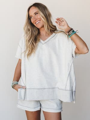 A relaxed matching short set in heathered light grey — oversized v-neck top with contrast stitching and a ribbed hem band paired with casual shorts, styled with stacked turquoise and beaded bracelets for that effortless, off-duty boho feel.