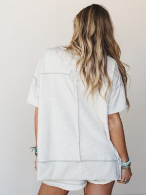 Back view of a relaxed heather grey oversized tee with exposed seam detailing, paired with cutoff shorts and layered turquoise beaded bracelets — the kind of easy outfit that looks effortless without trying.