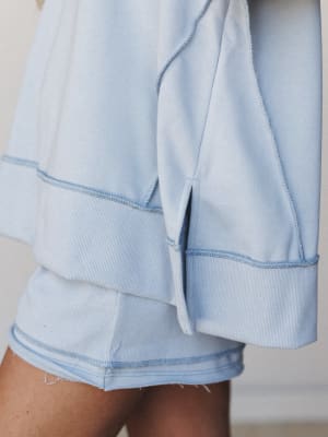 A close-up detail shot of a sky blue matching short set, highlighting the contrast stitching, ribbed cuff hem on the sleeve, and raw-edge shorts hem — all the cozy construction details that make this set feel intentional, not just thrown together.