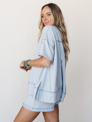 A soft sky blue oversized tee and matching shorts set styled from a side-back angle, accessorized with stacked turquoise and gold bracelets — the kind of effortless outfit that looks just as good on a coffee run as it does on the couch.