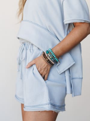 A close-up mid-shot of a sky blue matching short set — cropped top and drawstring shorts — styled with a stacked beaded bracelet set that adds just enough personality to keep the look from feeling too simple.