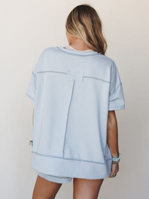 Shot from behind, a model wears a relaxed sky blue short-sleeve sweatshirt with visible contrast stitching and a back patch detail, paired with matching shorts — the kind of effortless set that makes staying in feel like a whole look.