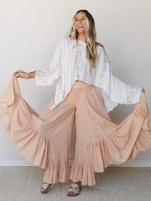 A laughing model fans out the dramatic ruffle hem of ultra-wide-leg pants in soft coral, paired with a flowy white embroidered top — the whole look feels like it belongs somewhere between a beach town market and a backyard dinner with good music.