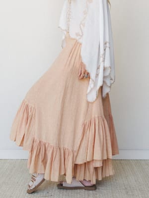 A dreamy side-profile shot of a tiered ruffle maxi skirt in soft peach-coral gauze, paired with a flowing white embroidered wrap top and strappy sandals — the whole look feels like a slow Sunday with nowhere to be.