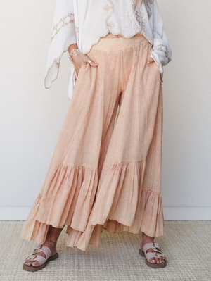 Flowy wide-leg pants in a soft peach-coral with ruffle hem details, styled with a white embroidered top and flat sandals — the kind of outfit that feels as easy as it looks.