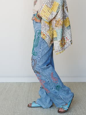 A side-profile shot of wide-leg denim jeans covered in colorful embroidered script detailing, styled with a patchwork kimono, turquoise beaded bracelets, and turquoise leather sandals — the kind of outfit that has a whole personality.