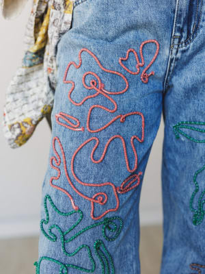 A close-up detail shot of medium-wash denim jeans covered in looping, raised embroidery in dusty rose and forest green — the kind of jeans that feel like wearable art and make you want to style them with something simple so they can do all the talkin
