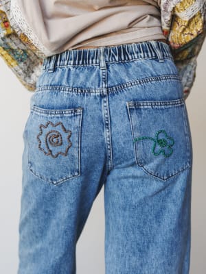 A close-up back view of light-wash wide-leg jeans with playful embroidered pocket details — a rust spiral sun on the left and a green clover on the right — styled with a floral ruffle jacket and a soft taupe top peeking out at the waist.
