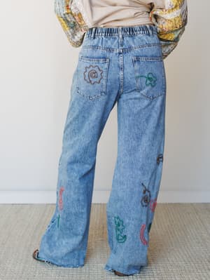Back view of wide-leg light wash jeans scattered with multicolor embroidered motifs — flowers, swirls, and abstract shapes — styled with a floral patchwork top for a playful, expressive boho look.