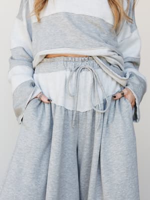 A close-up mid-shot of a relaxed matching set — wide-leg heather gray pants with a drawstring waist paired with a tie-dye sweatshirt in soft blue and cream, hands tucked in pockets with silver rings and dark nails adding a little personality.
