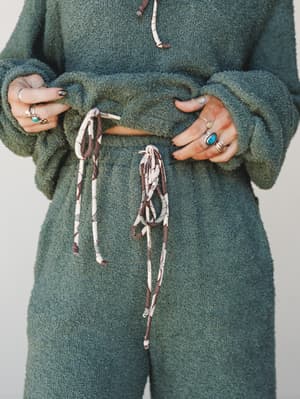 A mid-shot focused on the waist and hands of a model wearing a matching sage green boucle lounge set — cropped sweatshirt and wide-leg pants — styled with layered silver rings and a turquoise stone ring, holding the twisted fabric drawstring detail.