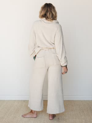 Shot from behind, a model in a matching cream boucle lounge set — cozy wide-leg pants and an oversized cowl-neck pullover — stands barefoot against a soft white wall, her turquoise jewelry the only pop of color in an otherwise cloud-soft look.