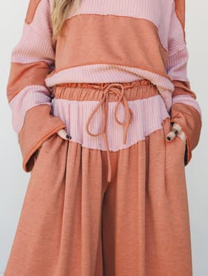 A close-up mid-shot of a color-blocked sweater and wide-leg pant set in warm camel and dusty mauve, with a waffle-knit layered waistband and drawstring detail — cozy and intentional in equal measure.