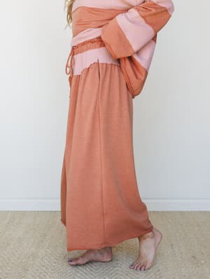 A side-profile shot of a relaxed wide-leg pant and color-blocked long-sleeve top set in warm camel and dusty pink, styled barefoot on a woven rug — easy, lived-in comfort with a cozy weekend feel.