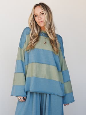 An oversized color-block pullover top in dusty teal and sage green with mixed fabric textures — part sweatshirt, part waffle knit — worn over a matching wide-leg pant in the same teal, styled with layered gold necklaces and stacked rings for that eff