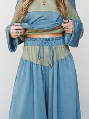 A close mid-shot of a layered two-piece set — a sage green waffle-knit crop top tucked under a dusty teal long-sleeve top, paired with flowy wide-leg pants in matching teal, all tied together with a drawstring waist and stacked boho rings.