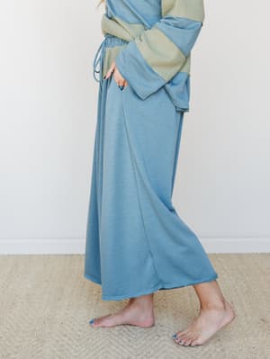 A side-angle mid-shot of a wide-leg maxi pant in dusty teal paired with a sage waffle-knit top, styled barefoot on a jute rug — the whole look has that intentionally relaxed, staying-in-but-make-it-pretty energy.