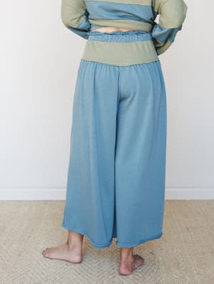 A relaxed two-piece set shot from the back — wide-leg crop pants in dusty teal paired with a sage waffle-knit top, worn barefoot on a woven rug against a soft white wall.