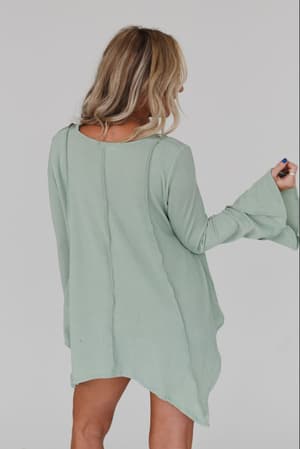 A blonde woman wears the Golden Hour Long Bell Sleeve Top in sage, standing against a plain white background. She faces away, glancing to the side, with her right arm bent and hand raised.