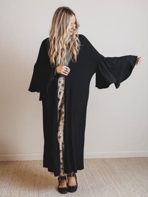 Woman wearing a long black duster over a patterned dress against a plain background