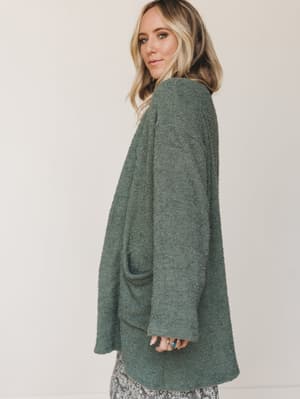 A soft, oversized boucle cardigan in muted sage green worn slightly off-angle — the kind of layer that makes you reach for it every single morning without thinking twice.