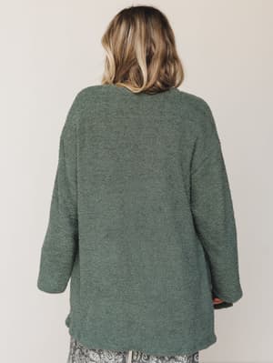 Shot from behind, a model wears an oversized bouclé cardigan in a muted sage-meets-forest green, paired with paisley-print pants — the whole look reads like a cozy Sunday that somehow got its act together.