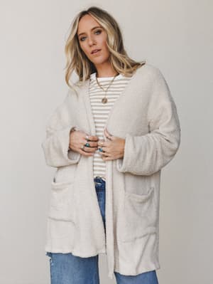 A cream boucle-textured cardigan worn open over a striped tee and distressed denim, styled with layered gold jewelry and turquoise rings — the kind of effortless layered look that makes you want to stay in it all day.