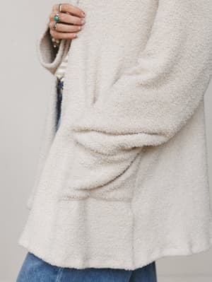 A close-up detail shot of a cream boucle cardigan layered over denim, with a hand full of stacked silver and turquoise rings peeking into frame — the kind of cozy you want to live in on a slow Saturday.