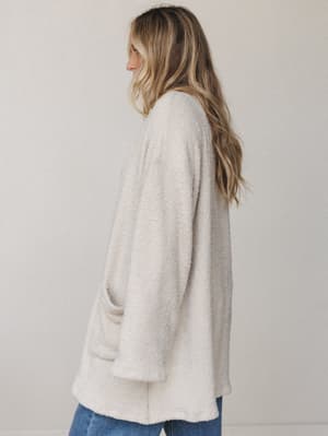 A soft, oversized boucle cardigan in a warm seashell tone, shot from a three-quarter back angle — the kind of sweater you reach for without thinking and never want to take off.
