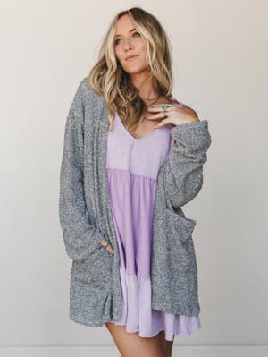 A relaxed, lived-in layering look — an oversized marled grey cardigan worn open over a lavender babydoll mini dress, styled with layered gold necklaces and a turquoise ring for that effortless boho-casual feel.
