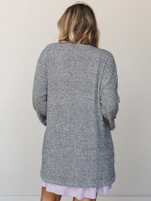 A back-view shot of a model in a heathered grey boucle-knit longline cardigan layered over a soft lavender mini dress, shot against a clean warm-white studio wall — cozy and collected without trying.