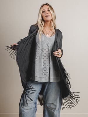 Woman wearing a black cardigan with fringe, gray top, and blue jeans against a beige background